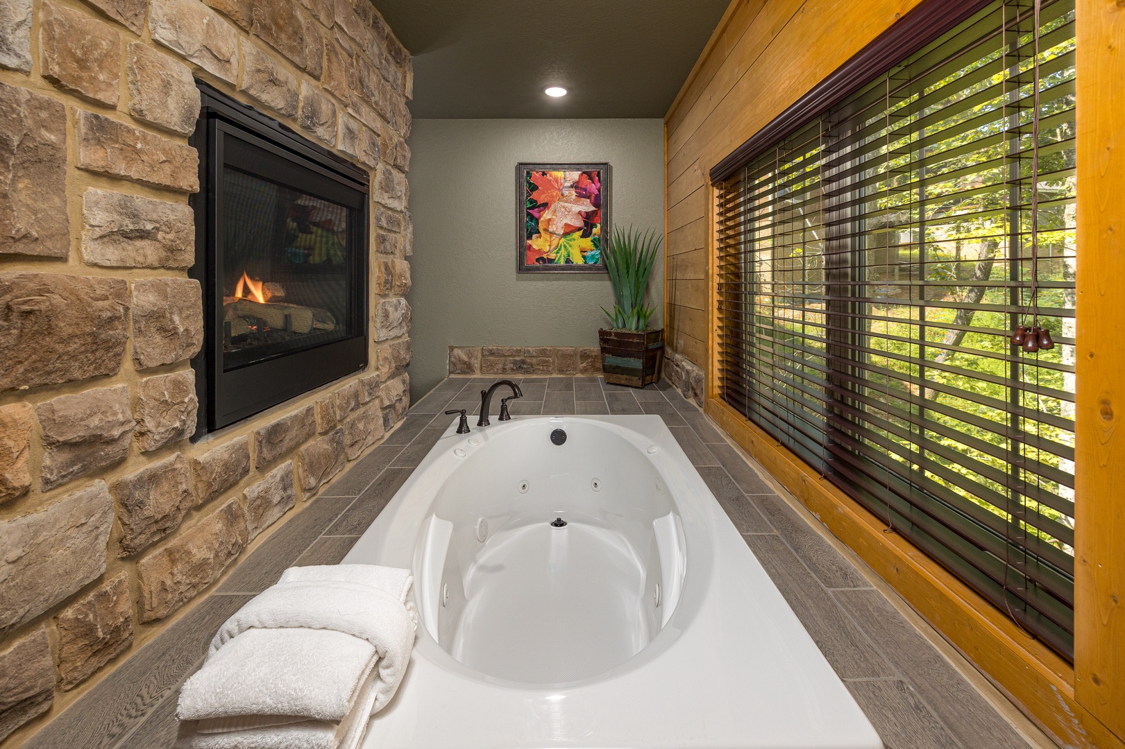 Southern Comfort spa bathroom — large round jetted soaking tub, stone fireplace, wood-panel accent wall, and garden window at Laurel Estates